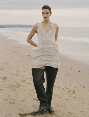 Model Beauïse Genç in a sleeveless knit tunic and black wader boots standing on a sandy beach for Numéro France.