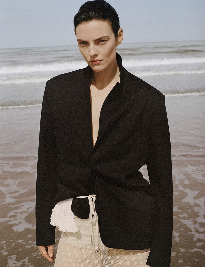 Beauïse Genç wearing a tailored black blazer and sheer skirt standing in shallow ocean water along the shore.