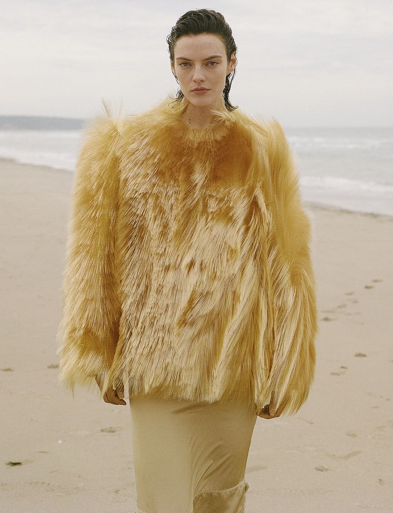 Model Beauïse Genç wearing an oversized textured golden cape top and beige skirt standing on a sandy beach.