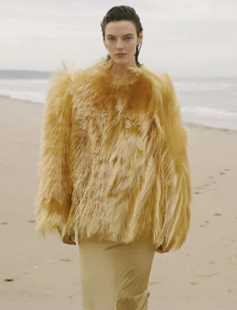 Model Beauïse Genç wearing an oversized textured golden cape top and beige skirt standing on a sandy beach.