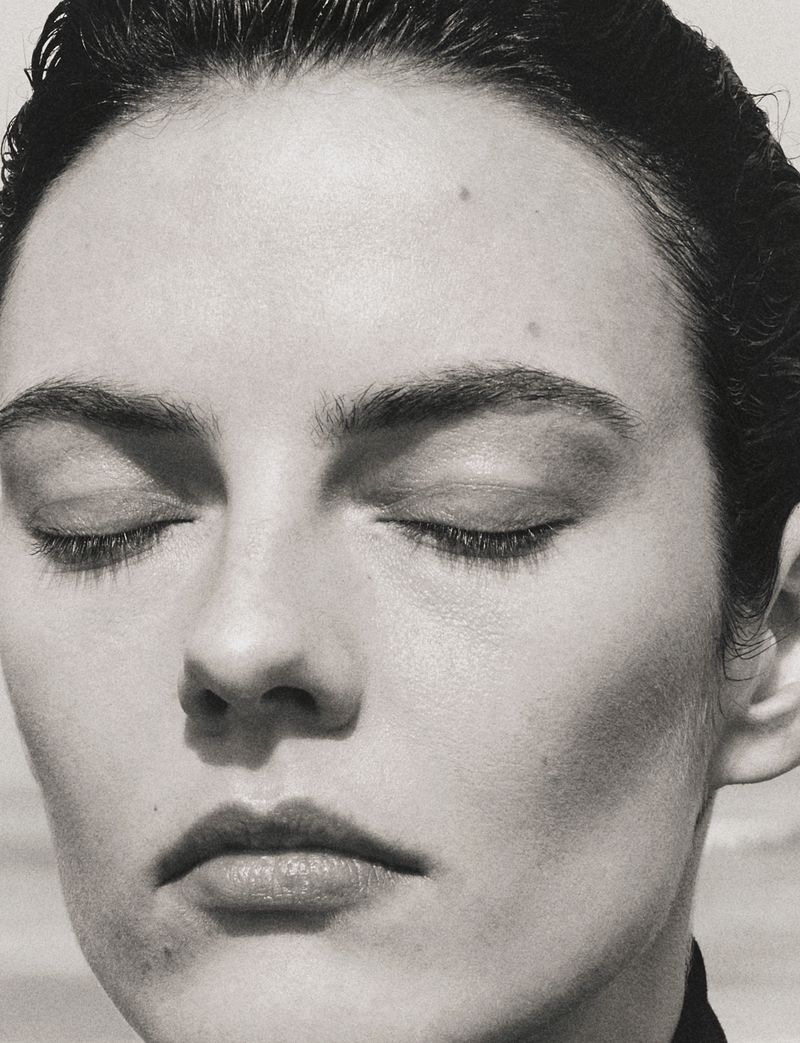 Close-up black and white portrait of model Beauïse Genç with eyes closed and slicked-back hair in bright natural light.