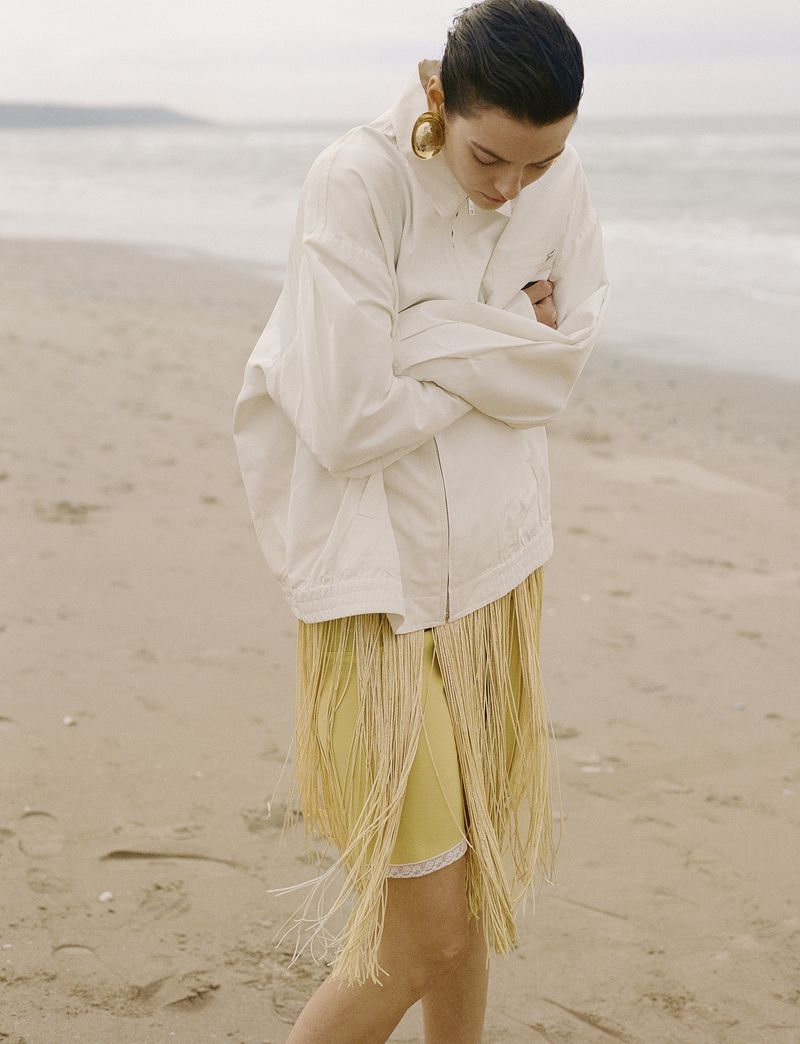 Model Beauïse Genç wearing an off-white jacket and yellow fringe skirt standing on a sandy beach looking down with arms folded.