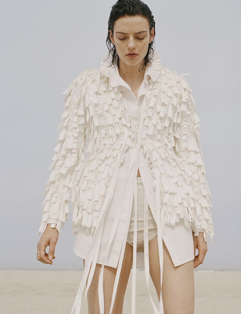 Model Beauïse Genç wearing a textured white fringed jacket with trailing ribbons against a pale neutral backdrop.