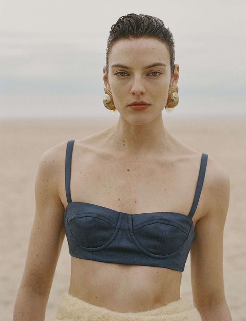 Model Beauïse Genç wearing a dark blue bustier top and gold shell earrings on a sandy beach for Numéro France.