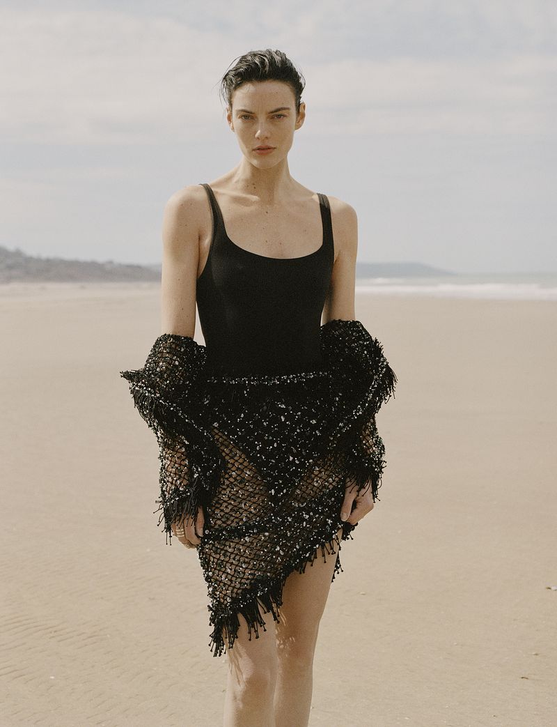 Model Beauïse Genç posing on a sandy beach in a black swimsuit and textured knit wrap.