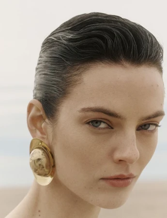 Close-up portrait of model Beauïse Genç with slicked-back dark hair wearing a large sculptural gold earring against a muted beach backdrop.