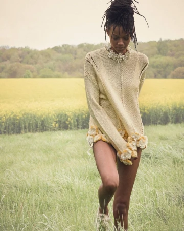 Model Nyela Hopster wearing a pale yellow knit sweater in a grassy field with a yellow blooming meadow in the background.