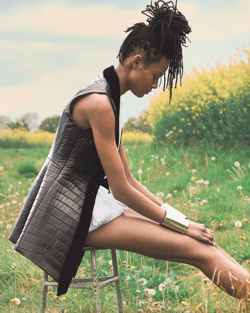 Model Nyela Hopster in profile wearing a textured sleeveless vest and silver cuff bracelet while seated in an open wildflower meadow.