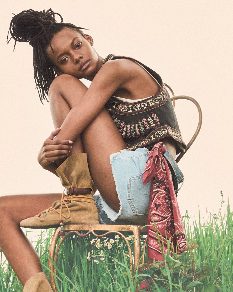 Model Nyela Hopster seated on a chair in tall grass wearing an embroidered vest, denim shorts, and tan suede boots.