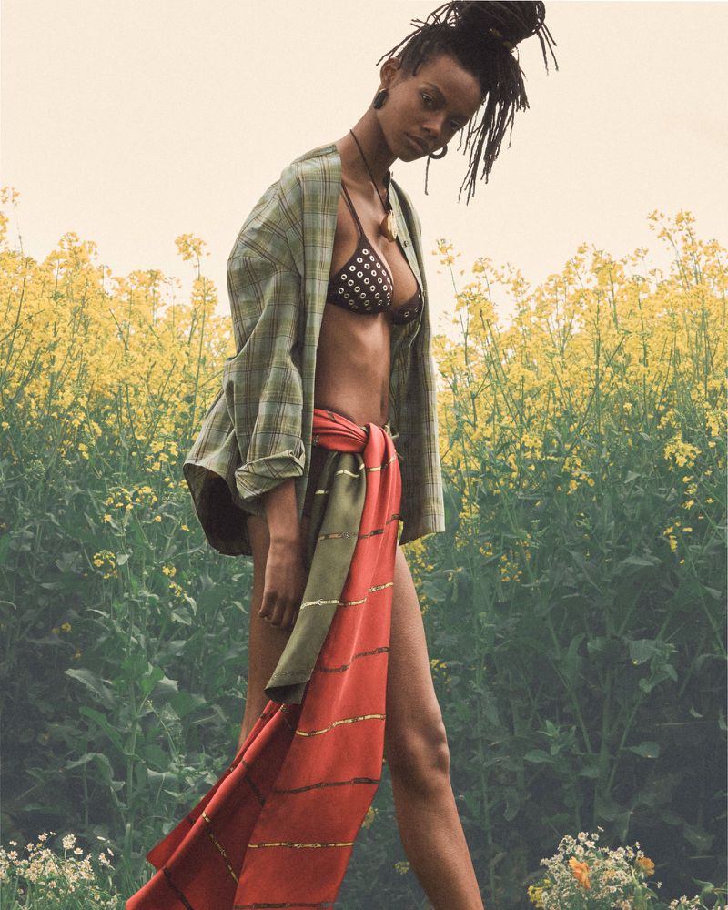 Model Nyela Hopster wearing a green plaid shirt, studded bikini top, and draped coral wrap skirt standing in a yellow wildflower field.