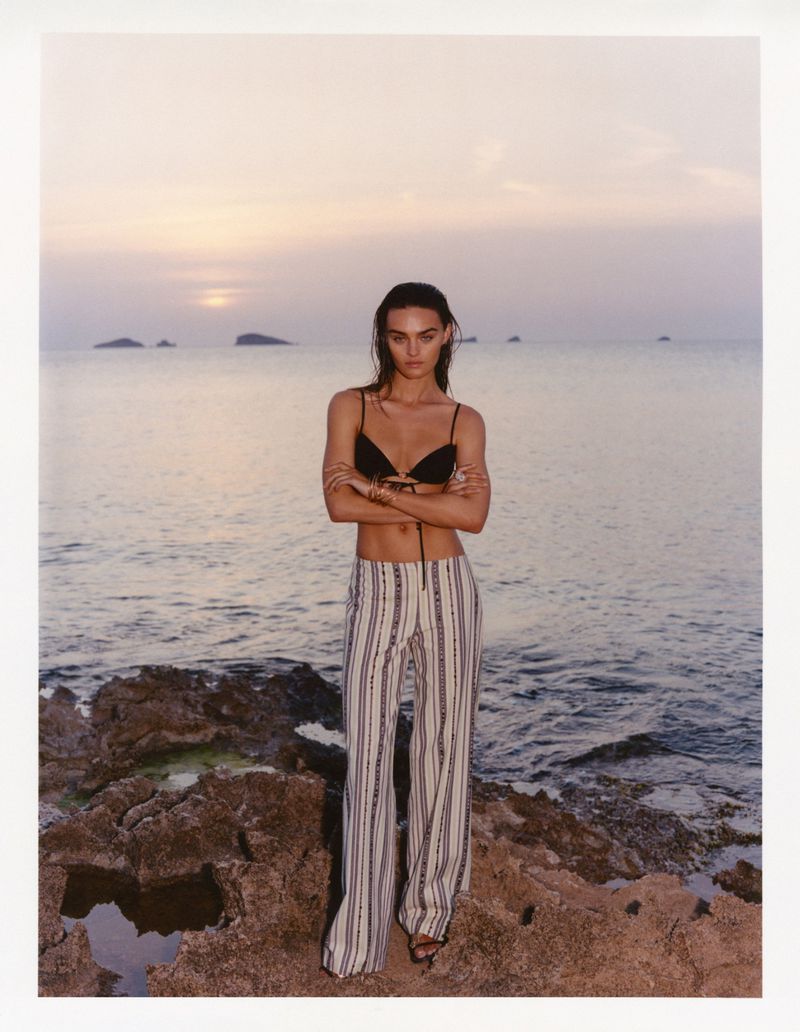 Model Anna Pricope in a black bikini top and striped trousers posing on a rocky coastline at sunset.