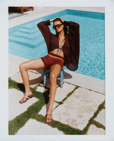 Model Anna Pricope sitting poolside in a dark brown tie-front blouse, knit shorts, sunglasses, and strappy sandals.
