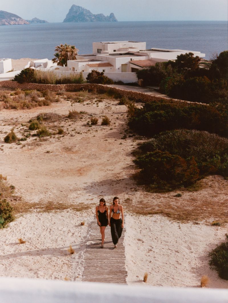 Two models walking together along a wooden boardwalk through a coastal landscape with white buildings and the sea in the background.