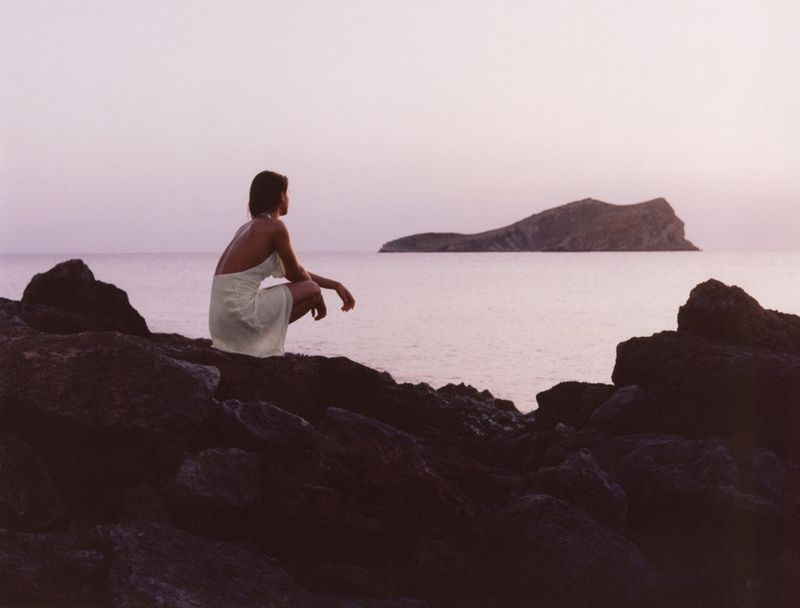 A woman in a white dress crouching on rocky shore overlooking a calm ocean with an island at sunset.