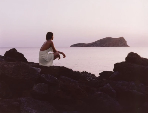 A woman in a white dress crouching on rocky shore overlooking a calm ocean with an island at sunset.