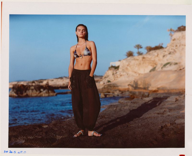 Model Anna Pricope poses on a rocky shoreline in a bikini top, black harem pants, and sandals for Pull & Bear Summer edit.