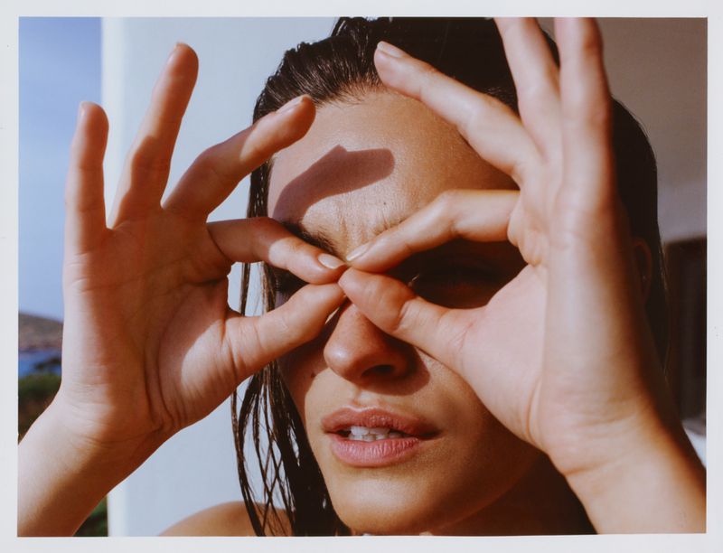 Close-up portrait of a woman looking through hand binoculars under bright direct sunlight with wet hair.