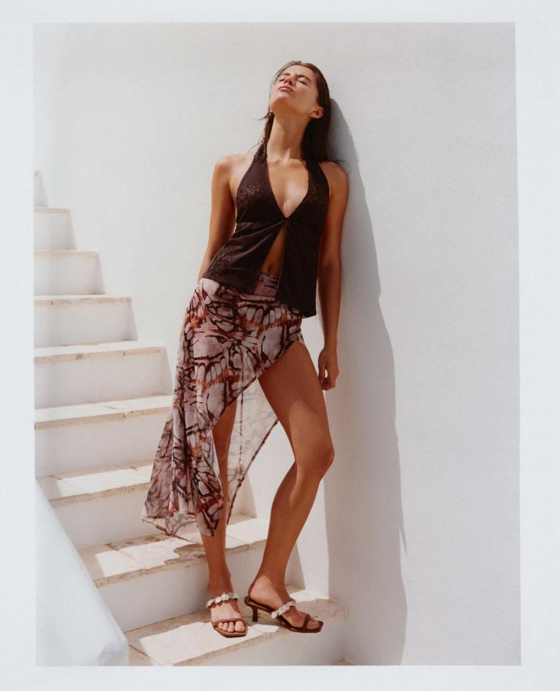 Model Anna Pricope in a dark halter top and sheer printed skirt leaning against a white wall by sunlit stairs.