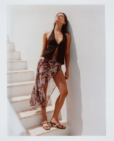 Model Anna Pricope in a dark halter top and sheer printed skirt leaning against a white wall by sunlit stairs.