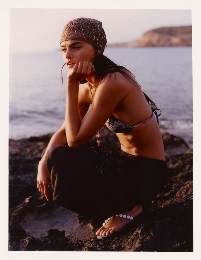 Model Anna Pricope crouching on rocky coast wearing a patterned headscarf and dark trousers at golden hour.