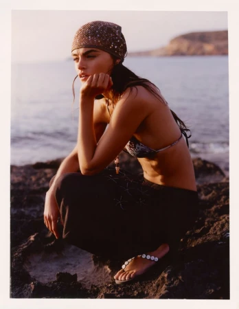 Model Anna Pricope crouching on rocky coast wearing a patterned headscarf and dark trousers at golden hour.