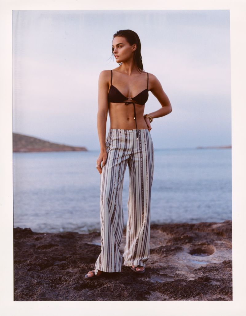 Model Anna Pricope wearing a black bikini top and striped wide-leg trousers standing on a rocky shoreline by the sea.