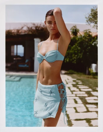 Model Anna Pricope poses beside a swimming pool wearing a light blue bikini top and a matching tie-up starfish mini skirt.