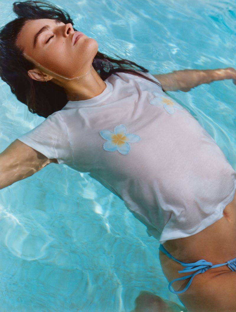 Woman floating on her back in turquoise pool water wearing a white graphic t-shirt and blue bikini bottom.