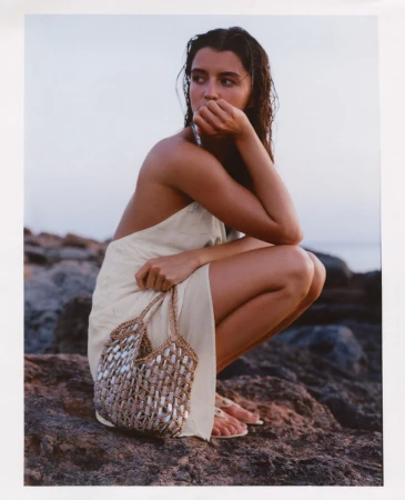 Model Anna Pricope crouching on a rocky coast wearing a cream halter dress and holding a woven mirror tote bag.