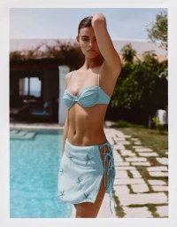Model Anna Pricope poses beside a swimming pool wearing a light blue bikini top and a matching tie-up starfish mini skirt.