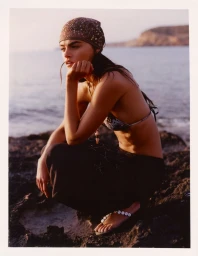 Model Anna Pricope crouching on rocky coast wearing a patterned headscarf and dark trousers at golden hour.