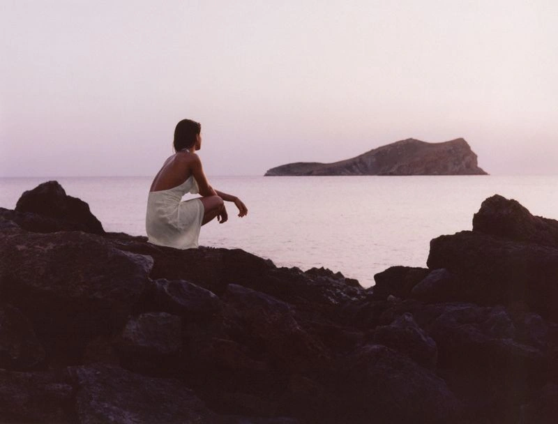 A woman in a white dress crouching on rocky shore overlooking a calm ocean with an island at sunset.