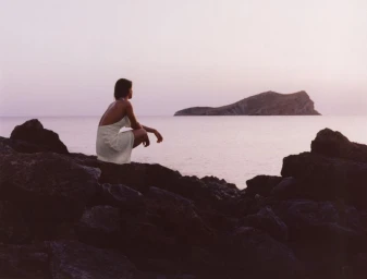 A woman in a white dress crouching on rocky shore overlooking a calm ocean with an island at sunset.
