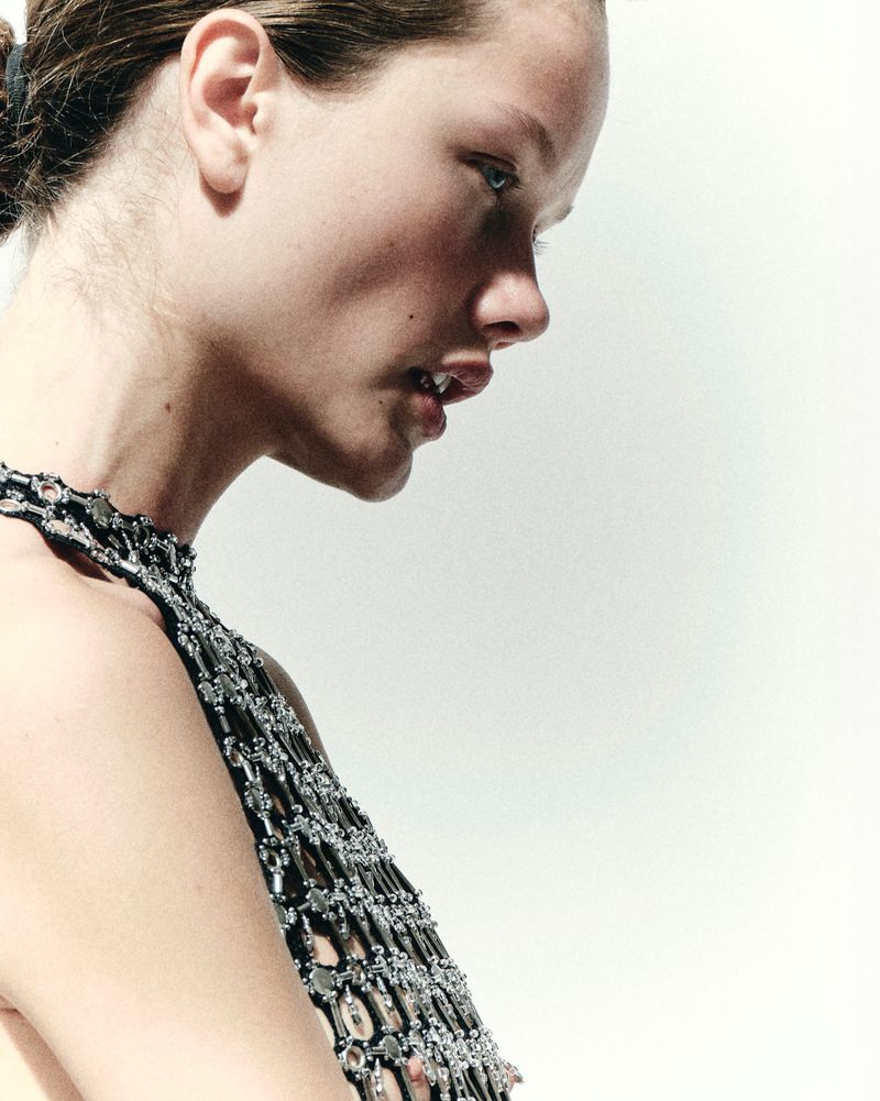 Close-up of a young adult woman in profile wearing a black beaded top against a white backdrop in a fashion or beauty context.