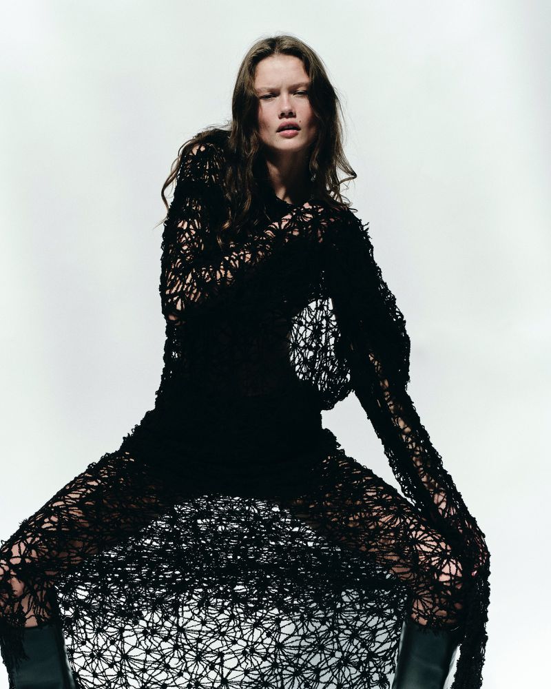 A woman with long brown hair wears a black textured net dress and black boots, sitting in front of a white background in a studio shot.