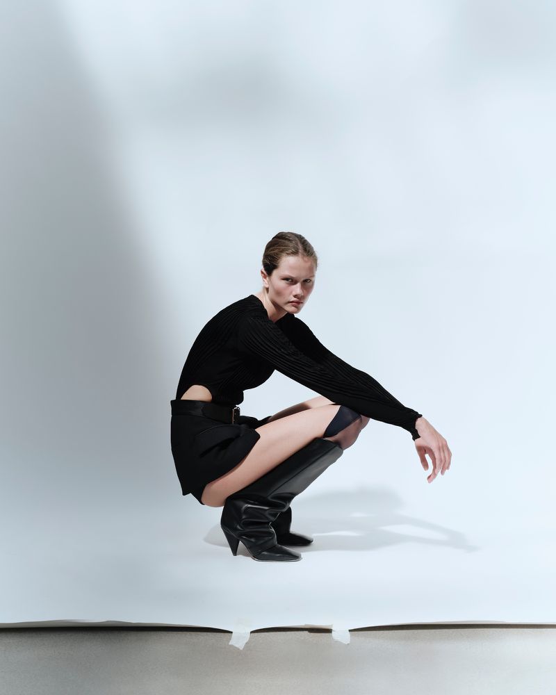 Fashionable woman squats in black cutout sweater, mini skirt, and knee-high boots against a white backdrop in a studio setting.