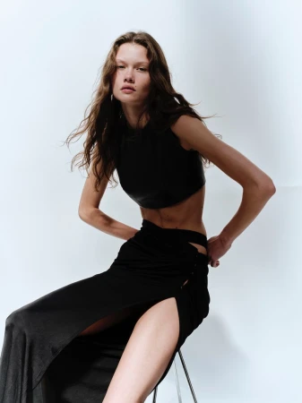 Model Tessa Jean seated wearing a black sleeveless crop top and high-slit skirt against a plain studio corner.