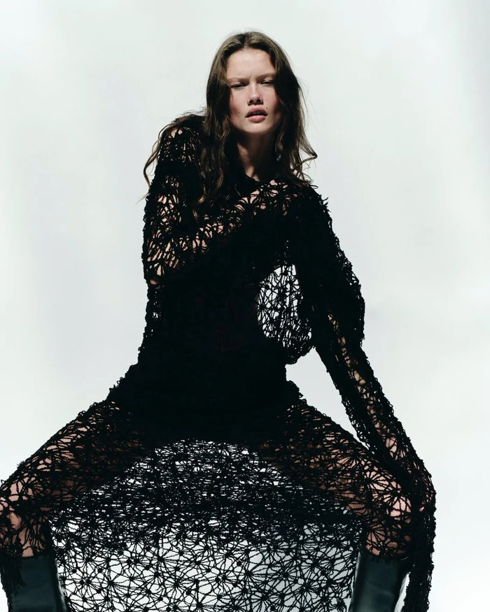 Model Tessa Jean in a textured black open-knit dress and boots posing against a white studio background.