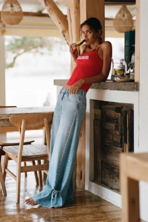 Anna Pricope wearing a red tank top and wide-leg light blue jeans leans against a rustic wooden bar counter.