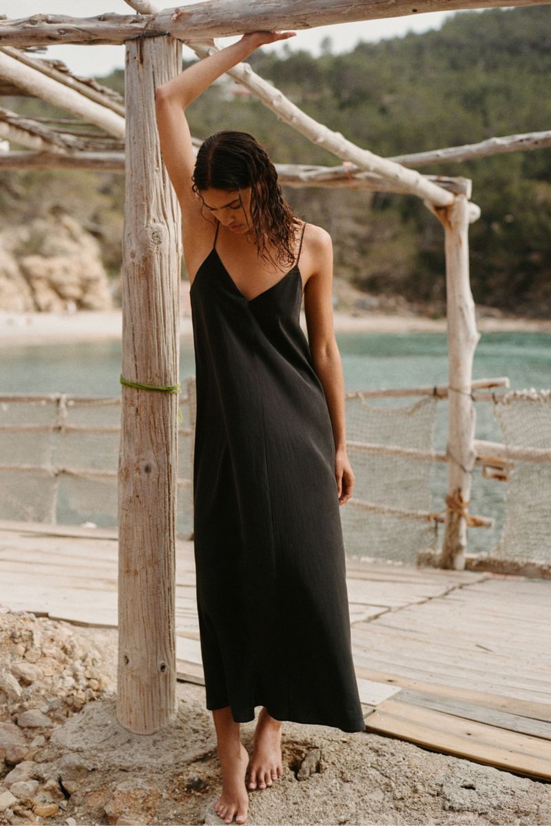Model Anna Pricope poses barefoot in a long black spaghetti-strap dress under a wooden pergola by the sea.