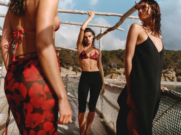 Three models wearing swimwear and summer apparel standing on a rustic wooden pier overlooking a coastal beach.