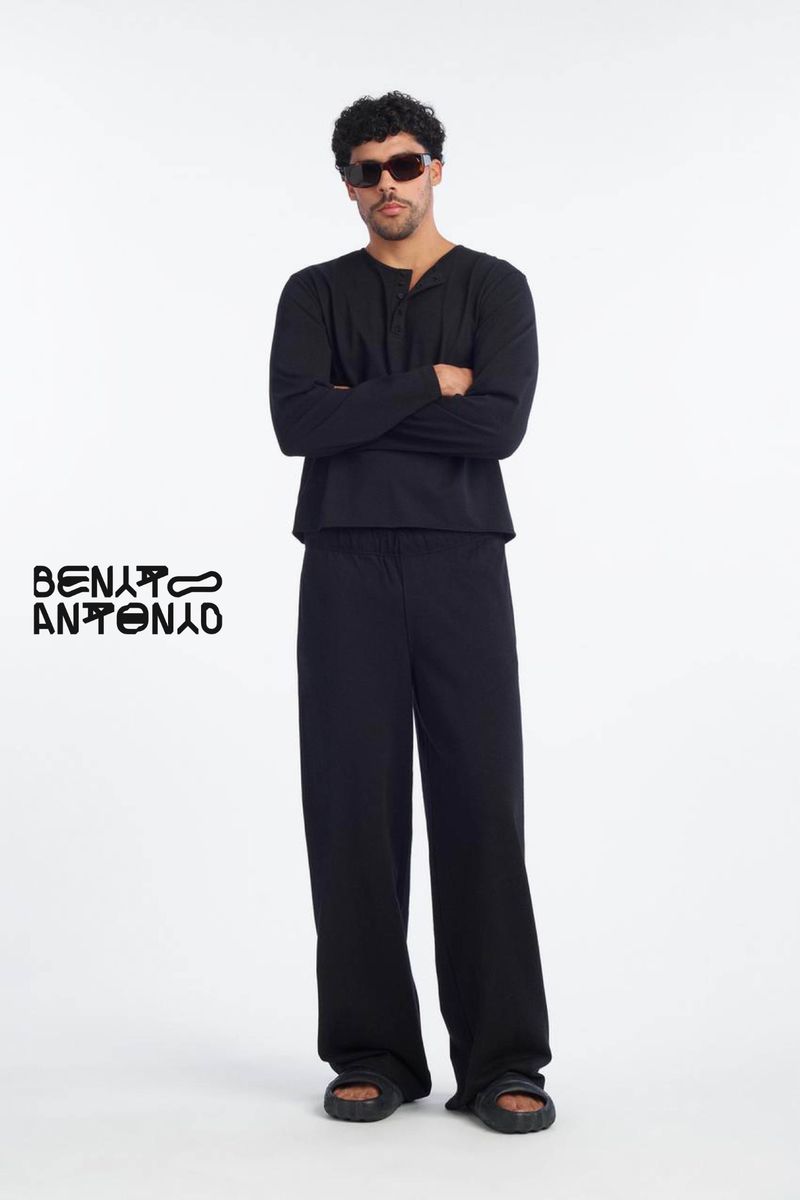 Man standing with arms crossed wearing black long sleeve henley shirt and black trousers against a plain white background