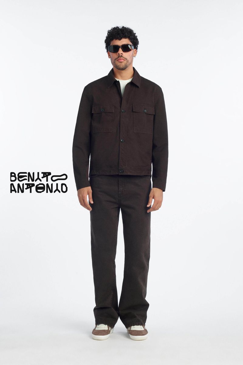 Model Bernie Martinez wearing a dark brown jacket and matching trousers with black sunglasses against a white studio background.