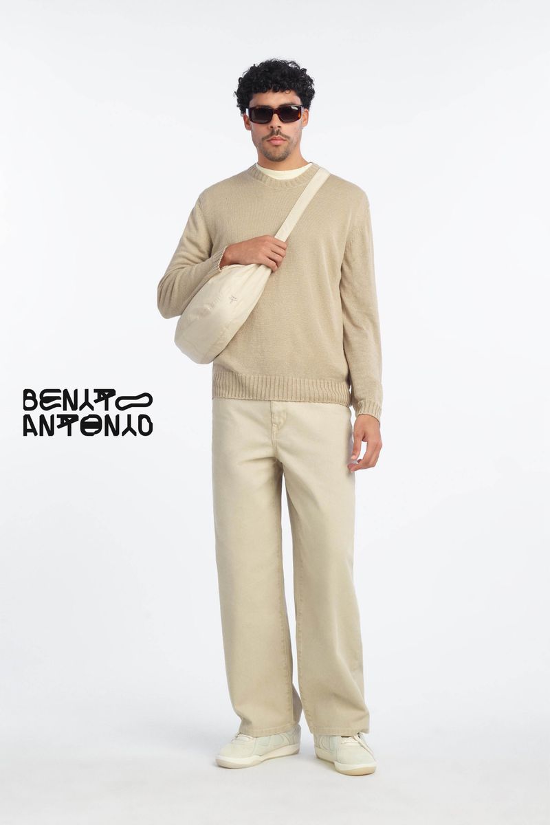 A man wearing sunglasses, a beige crewneck sweater, and trousers holds a crossbody bag against a plain white studio background.