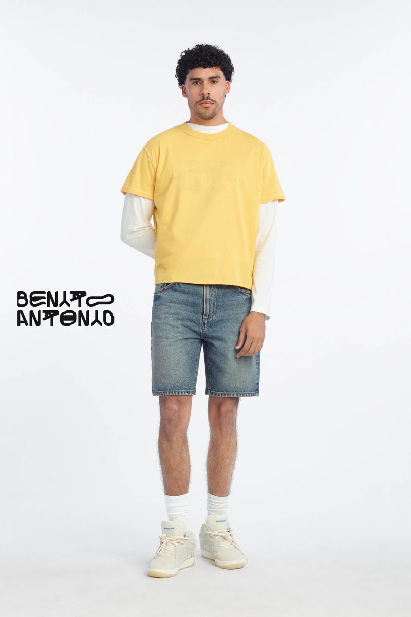 Man wearing a yellow t-shirt over white long sleeves and blue denim shorts against a plain white background
