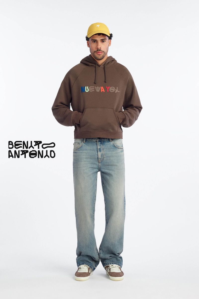 Bernie Martinez in a brown graphic hoodie, light-wash denim jeans, and a yellow cap against a white background for Zara.