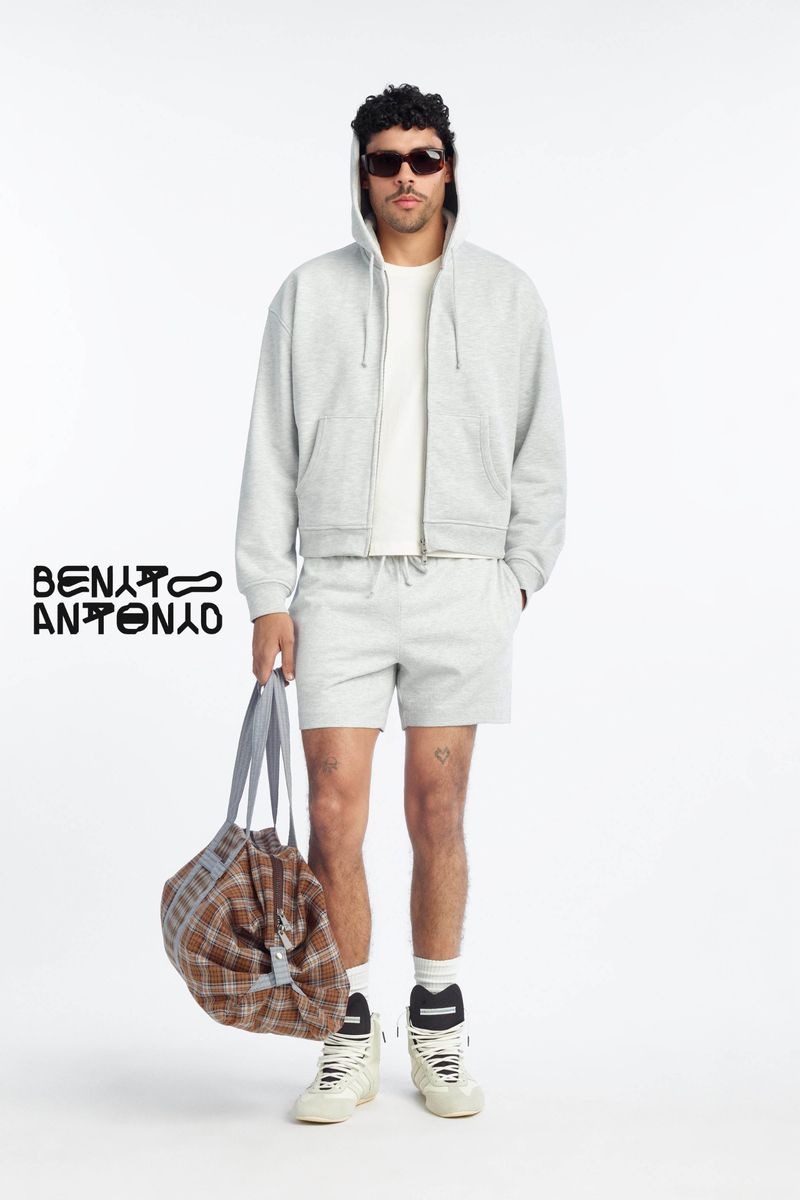 Model Bernie Martinez in a grey hoodie, shorts, sunglasses, and high-top sneakers holding a plaid duffle bag on a white backdrop.