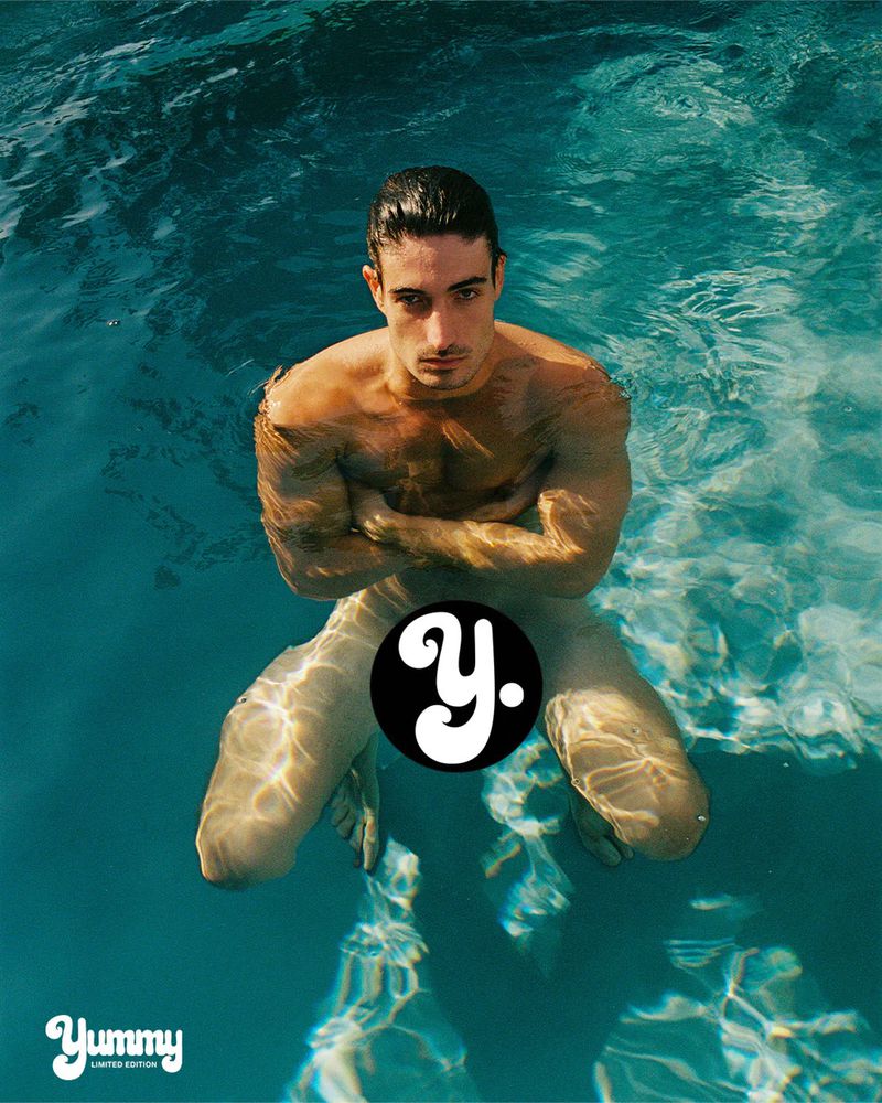 Model Romain Beziau partially submerged in clear turquoise swimming pool water looking directly at the camera in direct sunlight.