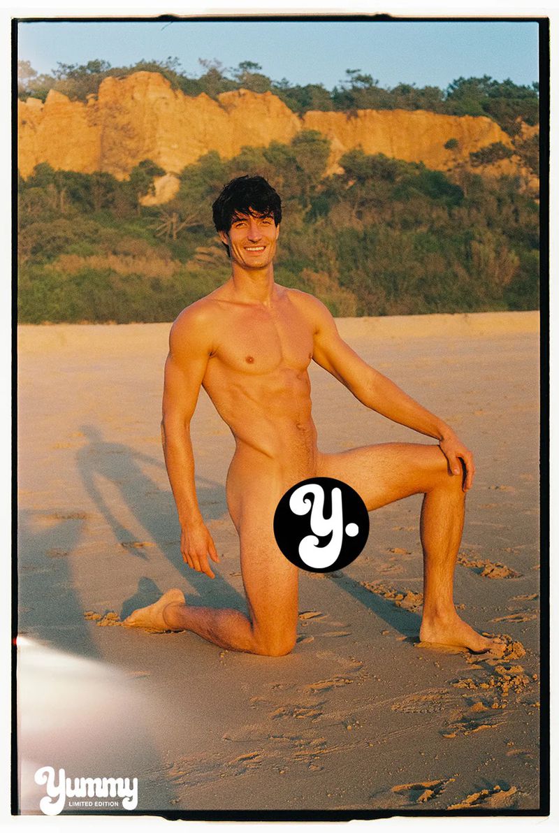 Model Romain Beziau kneeling on a sunny sandy beach with coastal cliffs visible in the background.