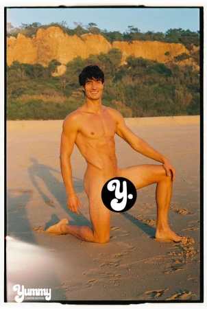 Model Romain Beziau kneeling on a sunny sandy beach with coastal cliffs visible in the background.